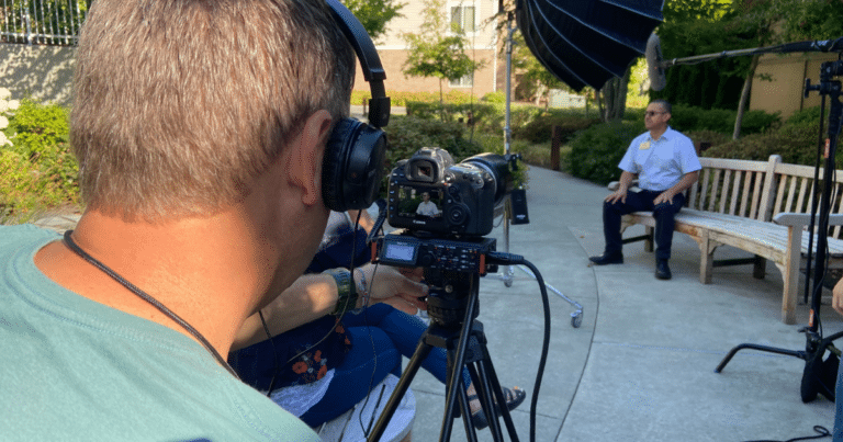 Photographer looking into a video camera while interviewing a man sitting on a bench.