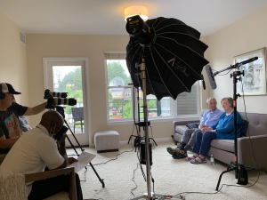 Behind the scenes of a husband and wife being interviewed with cameras and lighting equipment in front of them.