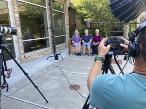 Looking over the shoulder of the cameraman interviewing 3 sisters sitting on a bench during a senior living photo shoot.