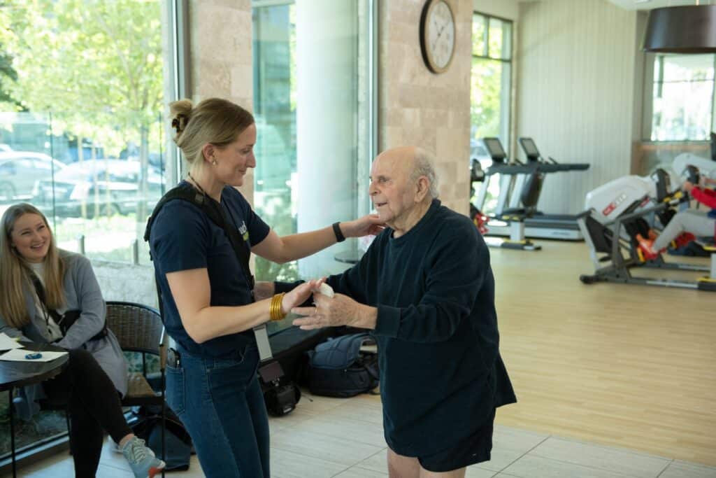 A young woman shakes hands with an elderly man in a blazer inside a gym, with exercise equipment and a seated person visible in the background.