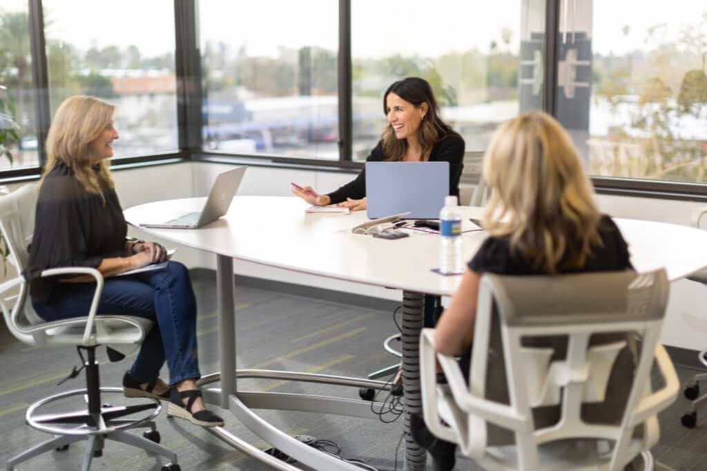 Three women sit around a white conference table in a bright office, each with a laptop as they discuss.