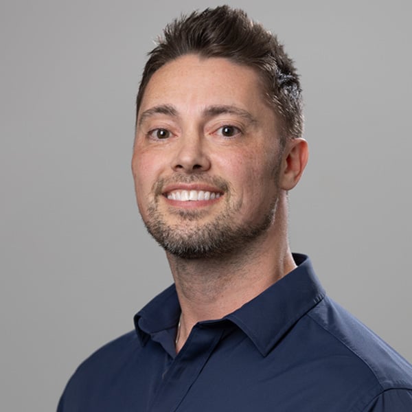 Portrait of a smiling man with short dark hair wearing a navy blue collared shirt against a gray background.