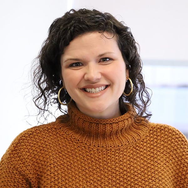 Smiling woman with dark, curly hair wearing a brown knitted turtleneck sweater and gold hoop earrings.