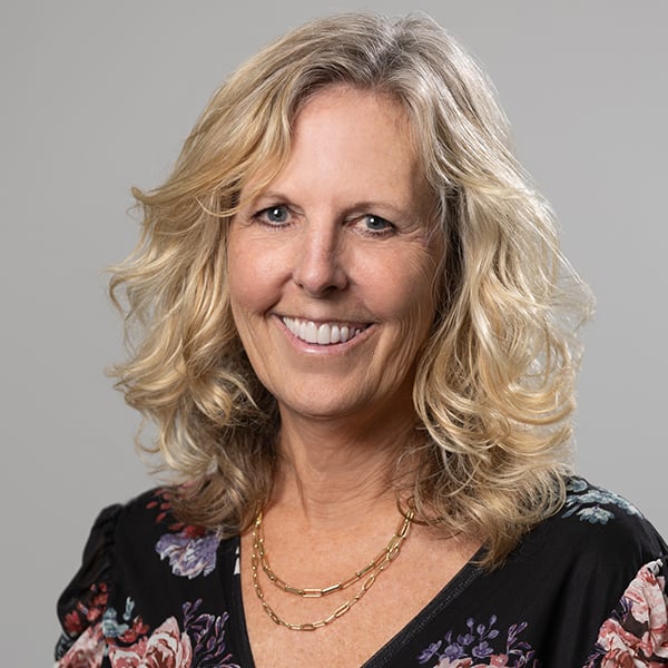 Smiling blonde woman with wavy hair wearing a black floral top and a layered gold necklace.