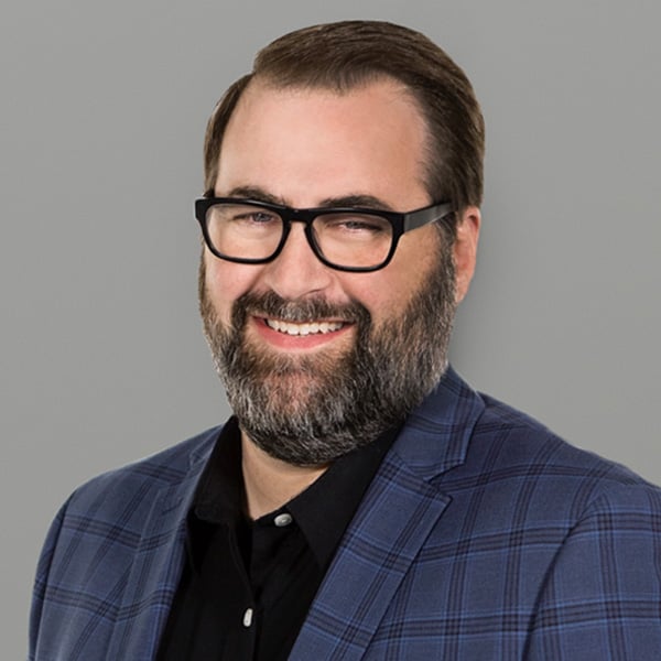 Smiling man with glasses and a full beard wearing a blue plaid blazer against a gray background.