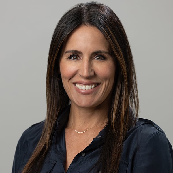 Portrait of a smiling woman with long dark hair wearing a navy blouse against a gray background.