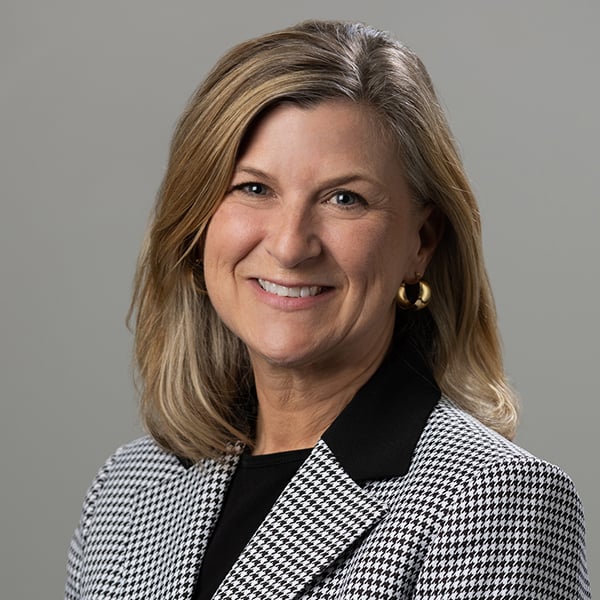 Smiling woman with blonde hair wearing a black-and-white checkered blazer and gold hoop earrings, posed against a gray background.