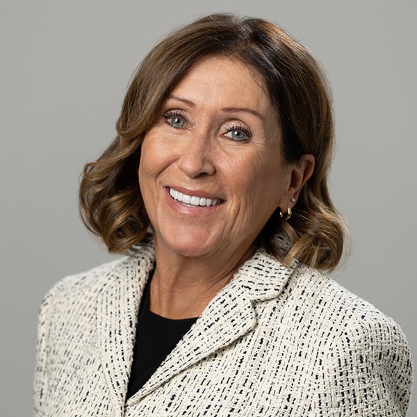 Smiling woman with short brown hair wearing a light patterned blazer against a gray background.