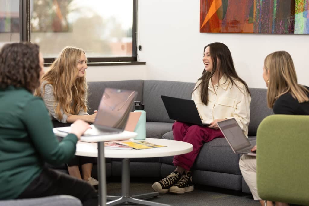 Four people sit around a round table with laptops, chatting in a bright office lounge.