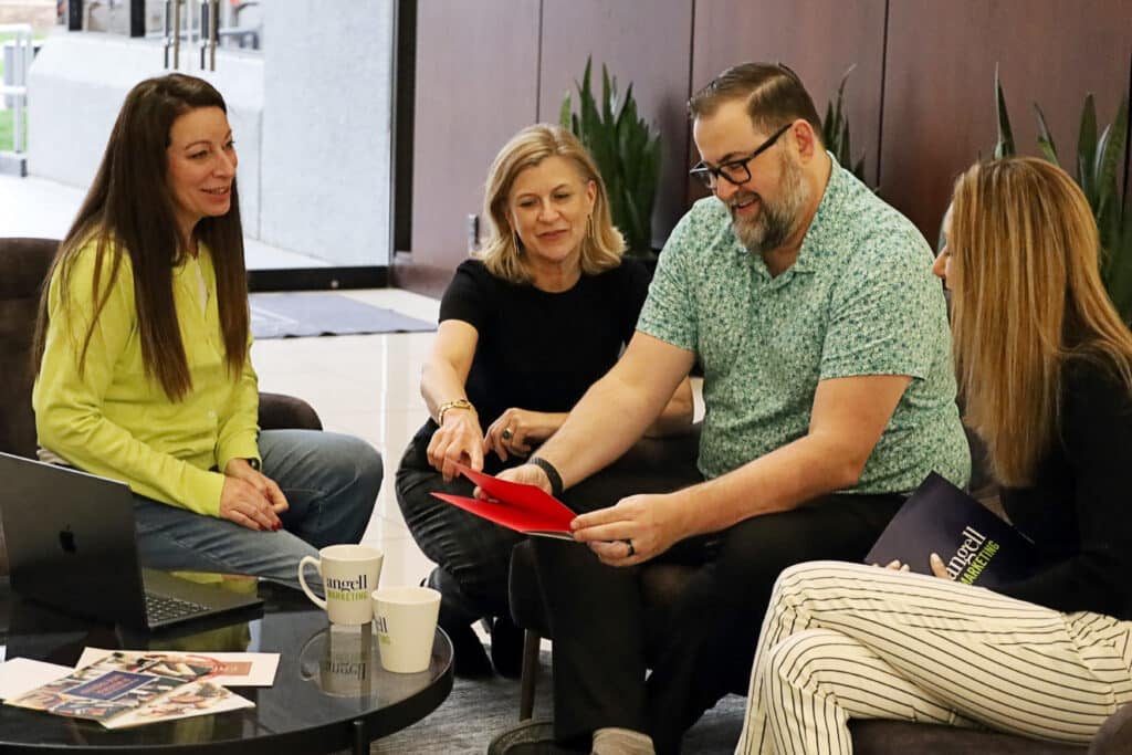 Four adults sit in a casual office lounge around a coffee table as a man in a green patterned shirt shows a red folder to the group.
