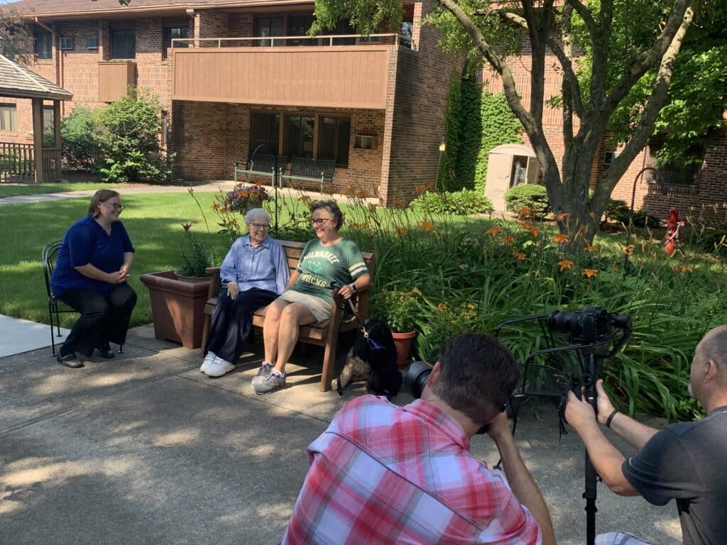 Two elderly people sit on chairs in a sunny garden in front of a brick building as two photographers film them.